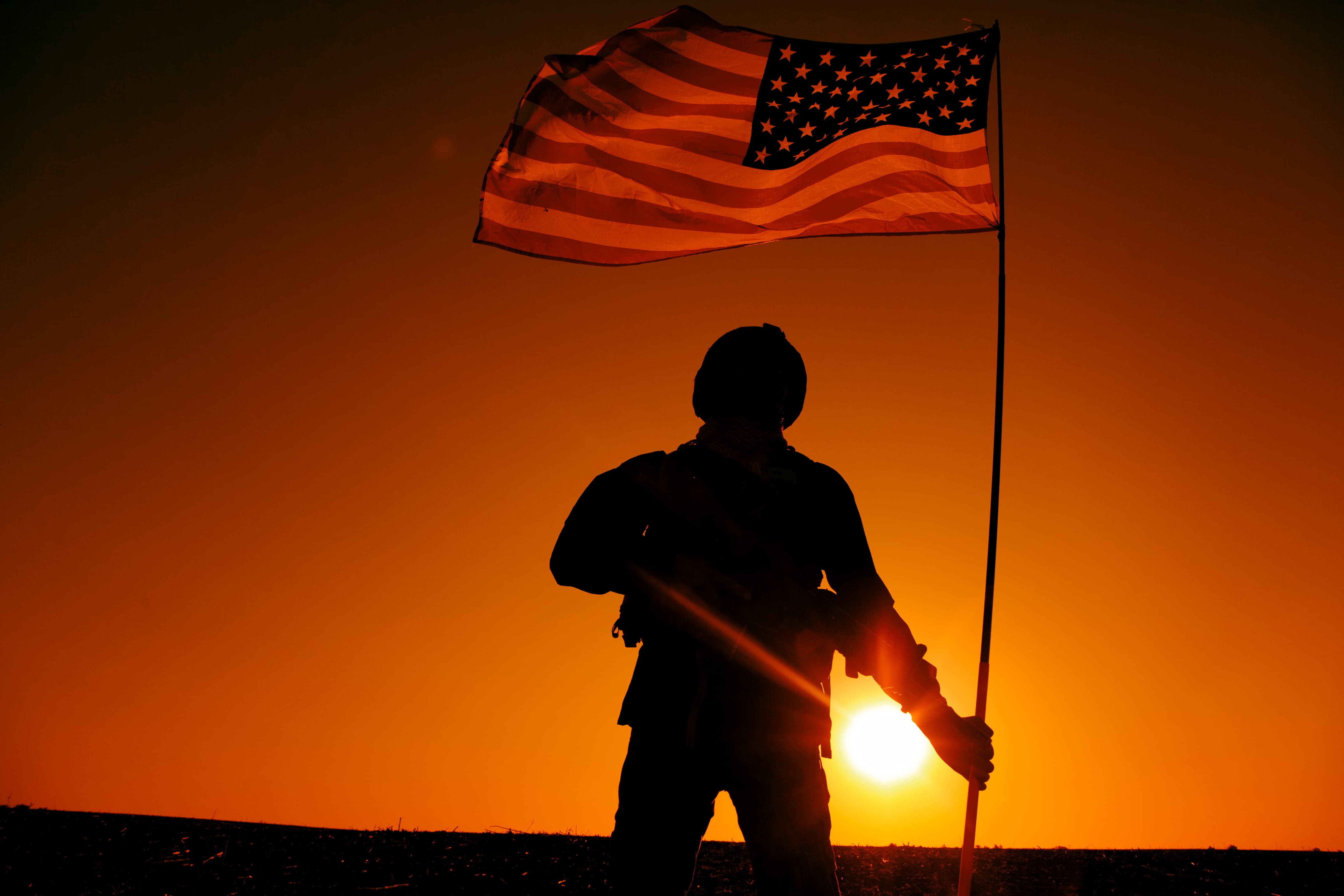 American Soldier silhouette over field with a background sunset while holding an American Flag high